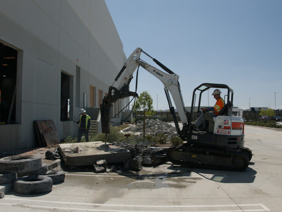 side view of technician in bobcat demolishing concrete slab from wall saw opening at ups facility in riverside, ca