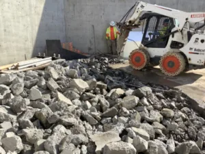 Side view of Bobcat with hydraulic breaker firing its chisel point into the concrete at Vans Skatepark in Costa Mesa