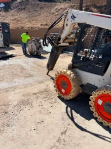 Intracut worker beside a flat saw watching a Bobcat loader operate a hydraulic breaker on concrete